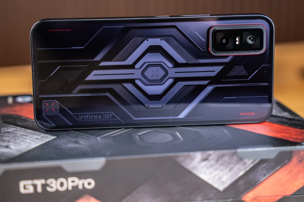 A horizontal product shot of the black Infinix GT 30 Pro smartphone, resting on its packaging. The back of the phone displays a detailed geometric pattern with a light-up feature. The 'Infinix GT' logo is visible, along with the camera module.