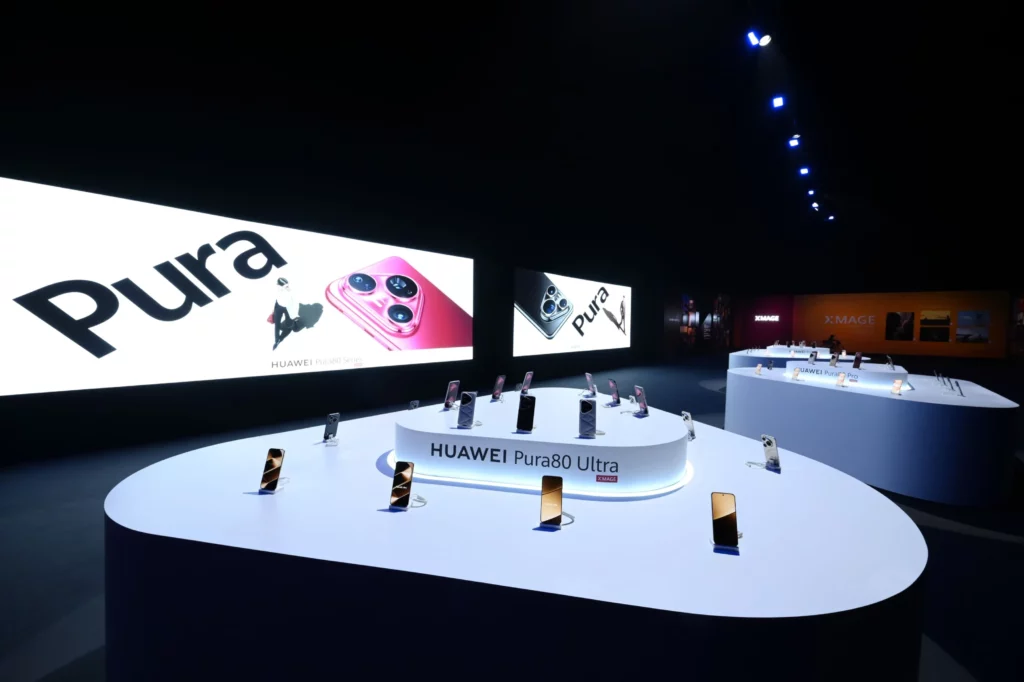 HUAWEI Pura 80 Series smartphones displayed on white pedestals in a dimly lit exhibition space, with large screens in the background showing 'Pura' branding and phone images.
