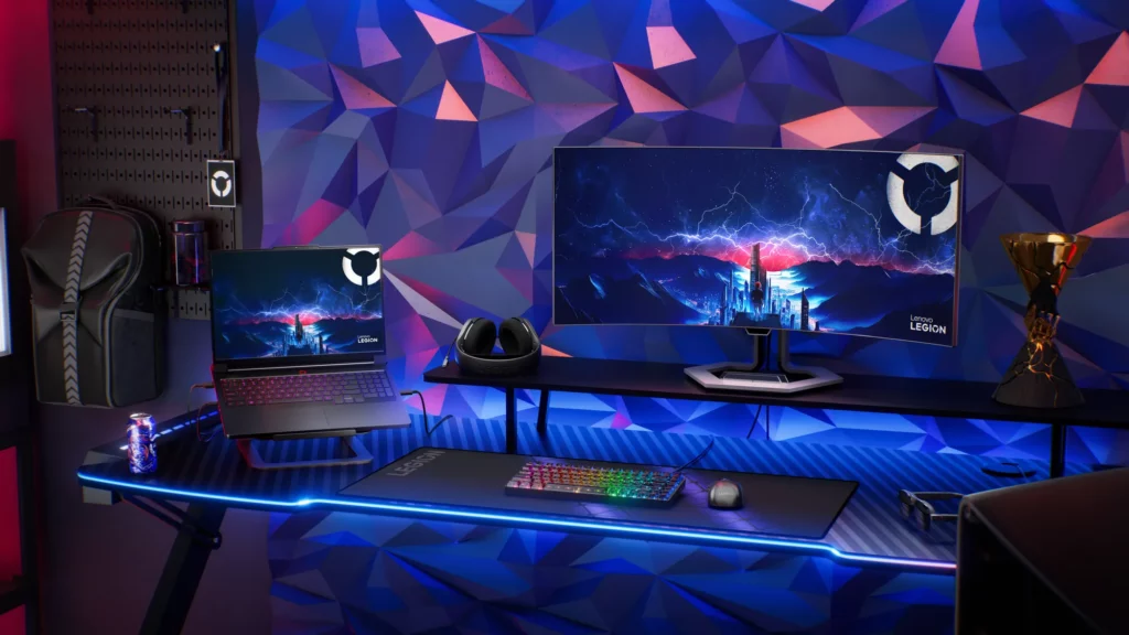 A brightly lit gaming setup featuring a Lenovo Legion gaming laptop, a large curved monitor displaying a game scene with the Legion logo, a colorful RGB keyboard, mouse, and headphones on a desk with blue LED lighting, all set against a vibrant geometric background
