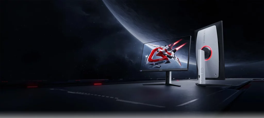 A Xiaomi Gaming Monitor G27i displayed on a dark, futuristic platform resembling a spaceship deck. The monitor is showing a dynamic image of a red and white mech-like figure in action. Another monitor is positioned upright to the right, showing its sleek, gray and black side profile with "Mini-LED" branding.