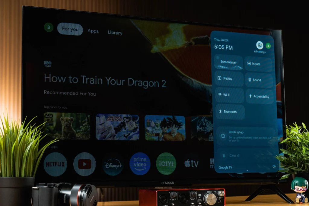 A smart TV displaying the Google TV interface. On the left is the main screen showing 'How to Train Your Dragon 2' as recommended content with app icons for Netflix, YouTube, Disney+, Prime Video, iQIYI, Apple TV, and HBO Max at the bottom. A blue overlay on the right shows the settings menu with options like 'Screensaver,' 'Inputs,' 'Display,' 'Sound,' 'Wi-Fi,' 'Bluetooth,' and 'Accessibility'.