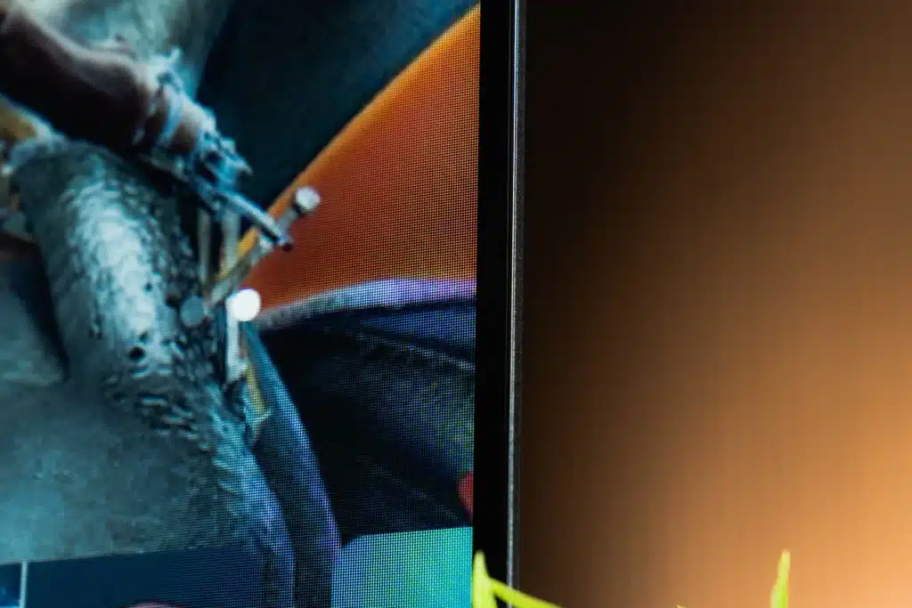 Extreme close-up of the side bezel of a television. The left half shows a pixelated image on the screen, while the right half shows the thin black side bezel and a blurred, warm-toned background.