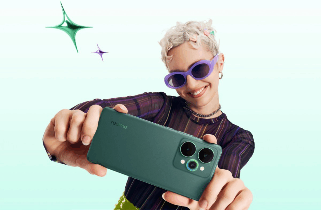 A person with short, light-colored curly hair and purple sunglasses holds up a teal green Realme smartphone to take a photo. The phone's back, featuring two large black camera circles and a smaller green lens, faces the viewer. The person is smiling broadly, wearing a dark striped top and a choker. Two stylized, shiny stars, one green and one purple, float in the light blue background.