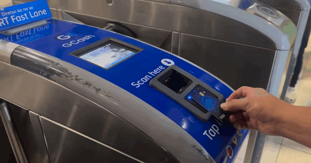 A close-up shot shows a person's hand tapping a blue GCash card on a blue and silver turnstile scanner. The turnstile has a screen displaying "GCash" and a prompt to "Scan here," along with a "Tap here" label near the card reader. The turnstile is metallic, with a sign partially visible in the background indicating a "Fast Lane."
