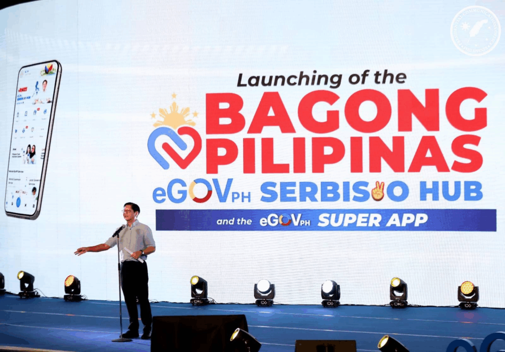 A stage setup for the "Launching of the Bagong Pilipinas eGovPH Serbisyo Hub and the eGovPH Super App." Philippine President Bongbong Marcos stands at a podium on the left, addressing an audience (not visible). A large screen behind him displays the event's title in prominent red and blue text with a stylized heart and crown. To the far left of the screen, a smartphone displaying a mobile app interface is also visible. Stage lights are arranged across the front of the stage.