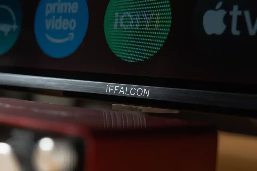 Close-up of the bottom bezel of a dark television displaying the white 'iFFALCON' logo. Above it, on the screen, are partially visible logos for Prime Video, iQIYI, and Apple TV.