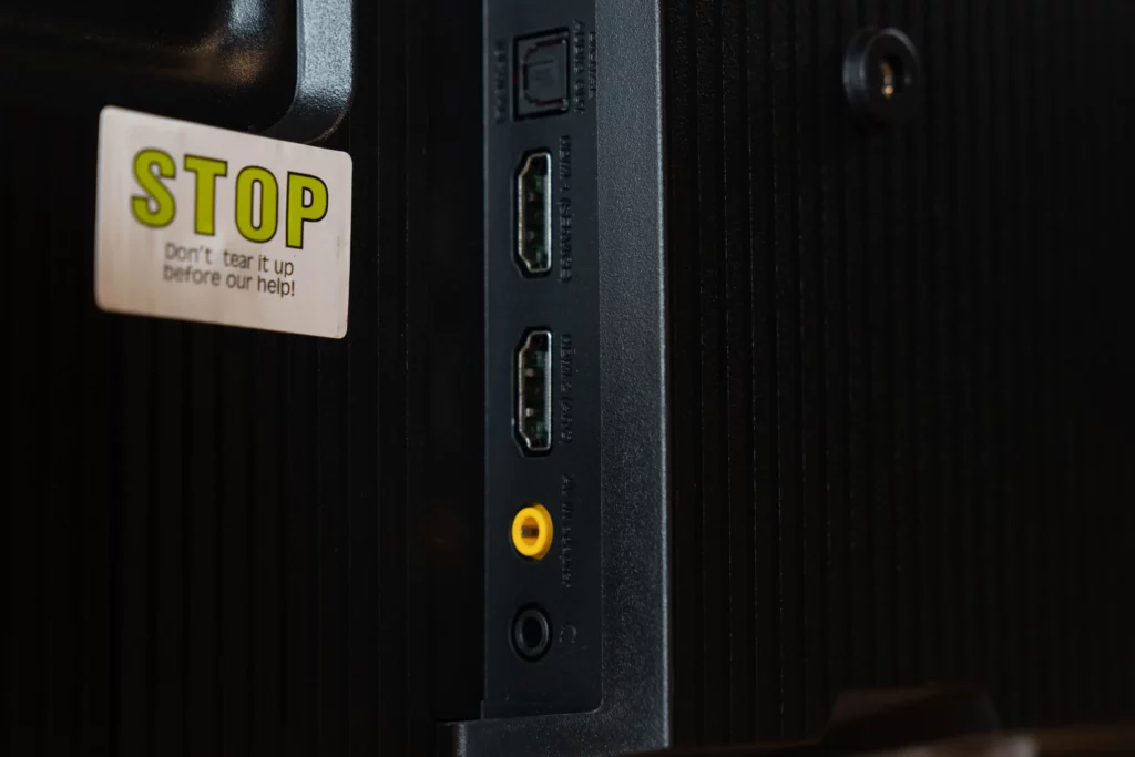Close-up of the back of a black television showing a yellow 'STOP' sticker on the left and several ports on the right. From top to bottom, there's an optical audio out, two HDMI ports, a yellow composite video input, and a 3.5mm audio jack.