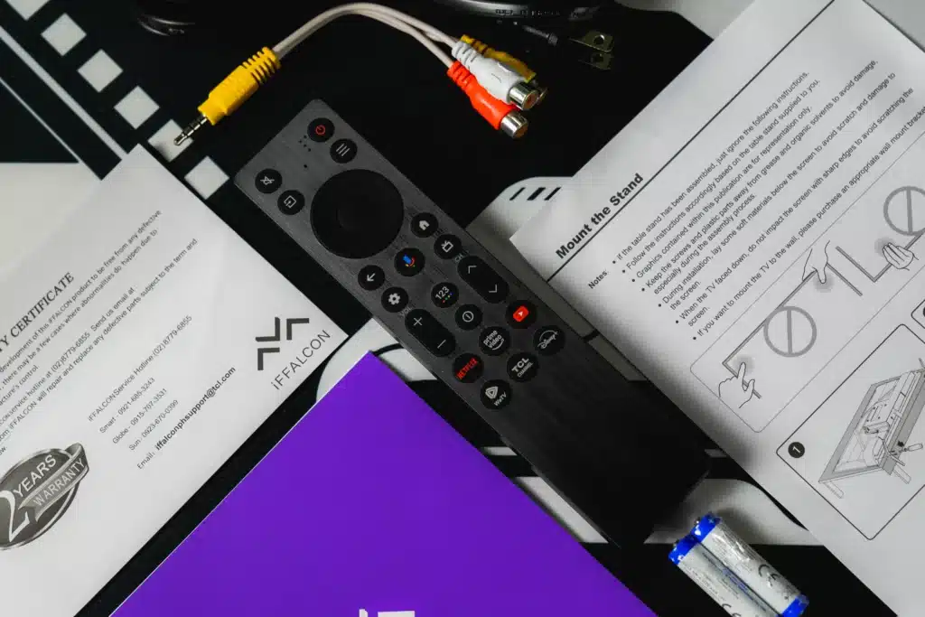 A black TV remote control with various buttons, including dedicated buttons for Netflix, Prime Video, and Disney+. The remote is resting on an open manual, with a yellow and white RCA cable and a red and white RCA cable in the background, along with two AAA batteries.