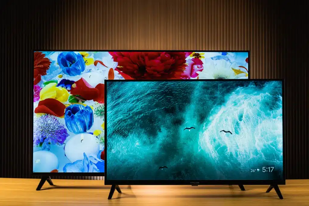 Two iFFALCON smart TVs of different sizes displayed side-by-side on a wooden surface. The larger TV in the background shows a vibrant image of colorful flowers. The smaller TV in the foreground displays a dynamic image of ocean waves with three birds flying and a time/temperature overlay.