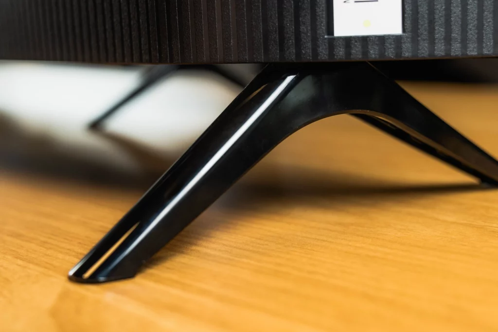 A close-up view of the shiny black plastic stand of a television, resting on a light brown wooden surface. The ribbed texture of the back of the TV is visible above the stand.