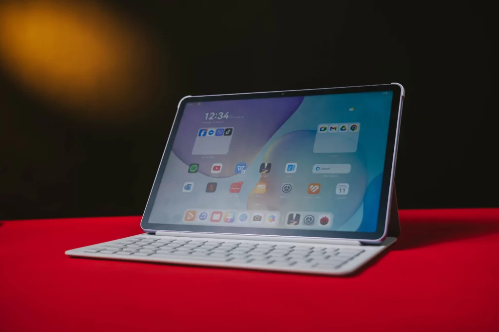 A purple HUAWEI MatePad 11.5 on a white smart keyboard, displaying the home screen with various apps like WPS Office, GoPaint, and Notes. The tablet is propped up on a red surface.