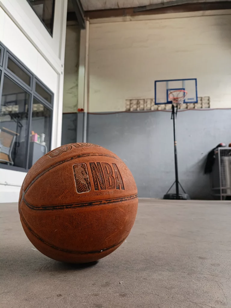 An NBA-branded basketball on the ground, with a basketball hoop visible in the blurred background.
