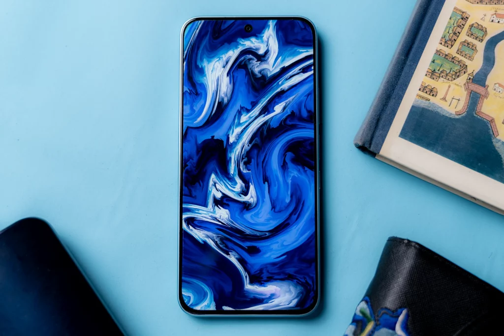 The front of the Infinix HOT 60 Pro displaying a dynamic blue and white abstract wallpaper, placed on a light blue surface next to a book and a wallet.