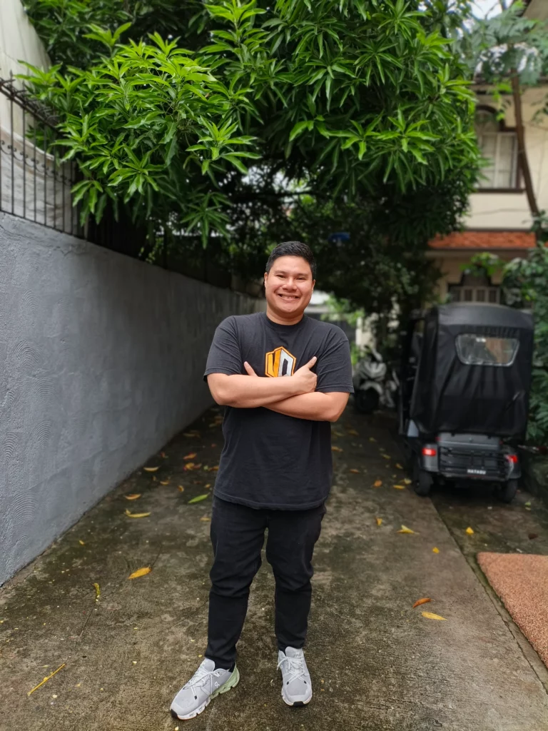 A man with his arms crossed, smiling, standing on a concrete path with lush green trees overhead.