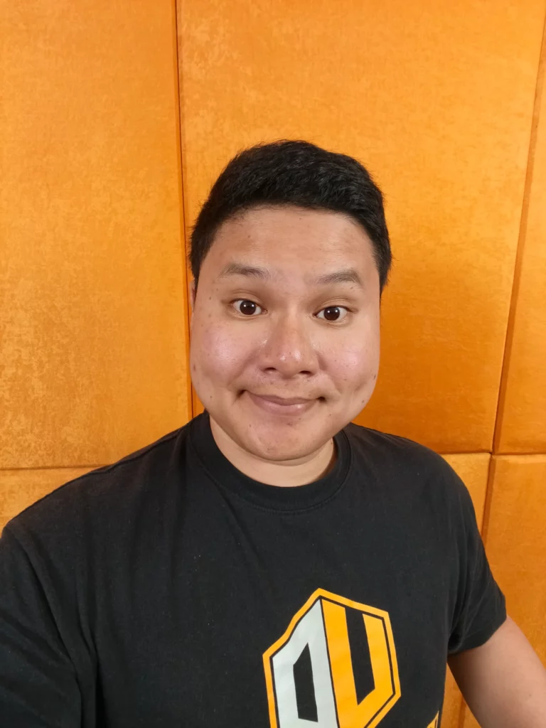 A man in a black T-shirt and glasses smiling at the camera, standing in front of an orange-paneled wall.