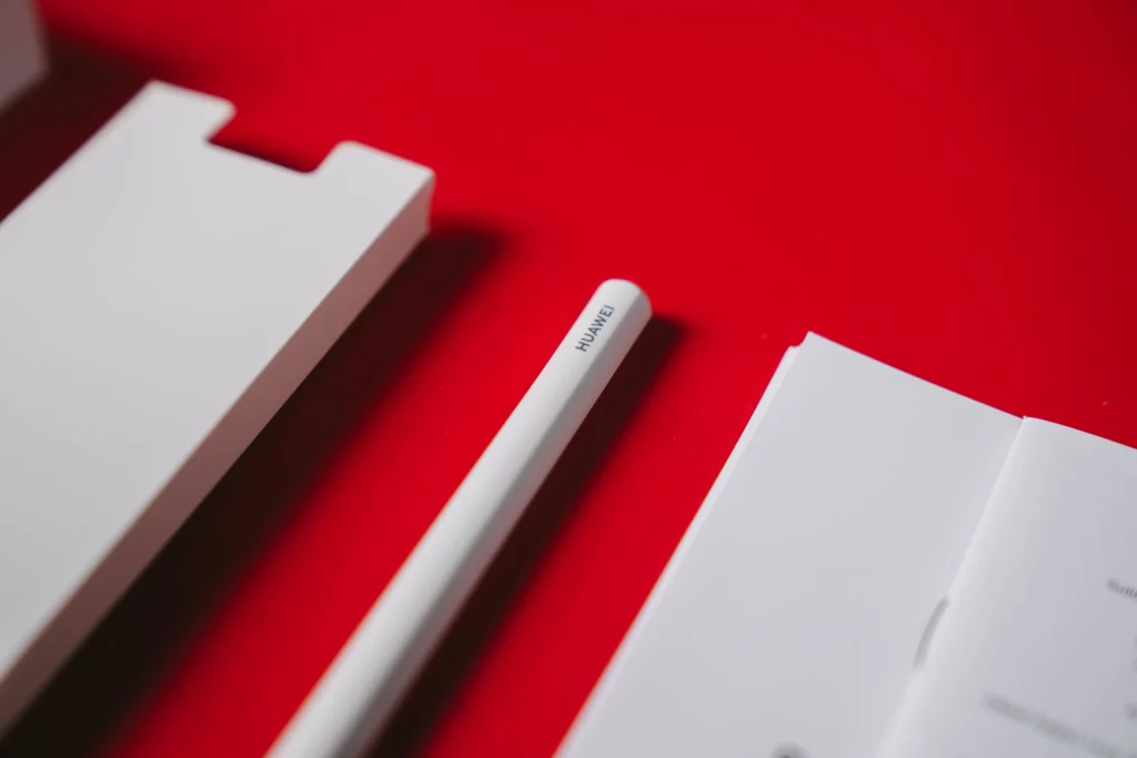 The white HUAWEI M-Pencil lying on a red background next to its packaging. The 'HUAWEI' logo is clearly visible on the side of the stylus.