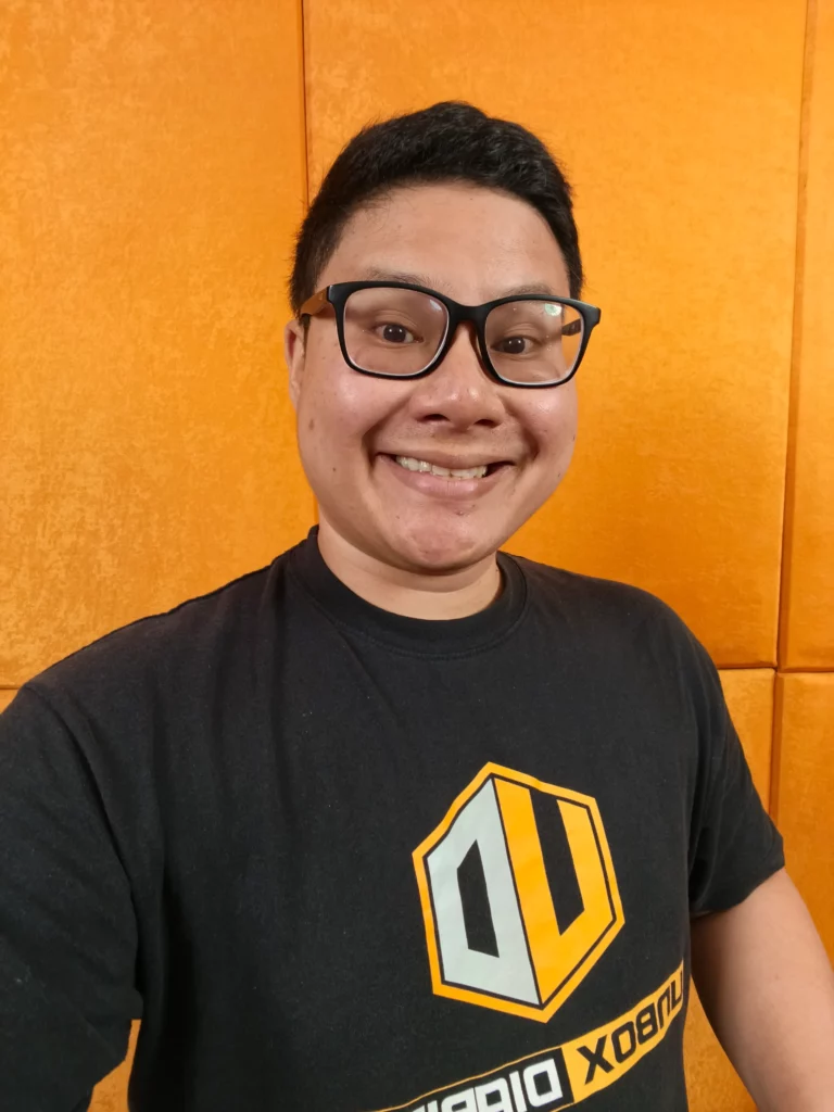 A portrait of a smiling man wearing glasses and a black T-shirt with a yellow logo, in front of an orange wall.