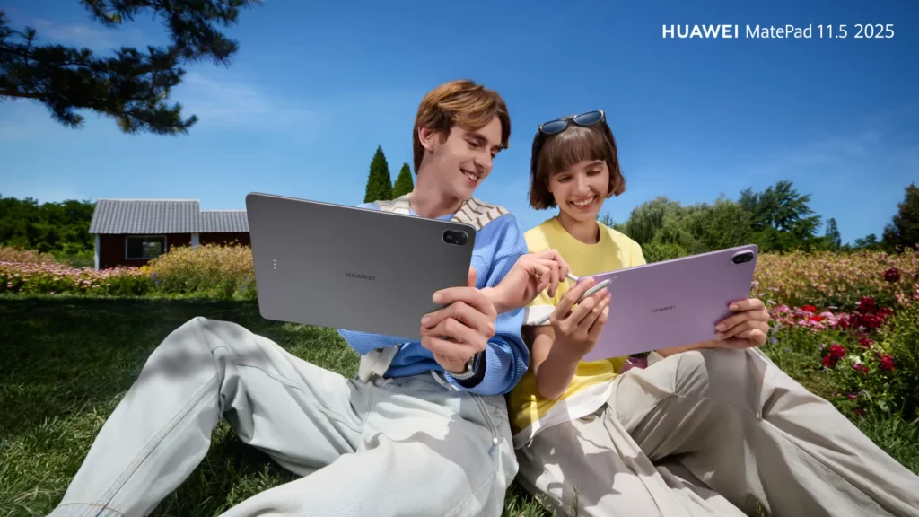 A young couple sits outdoors on a grassy hill, both using a Huawei MatePad 11.5 tablet in a serene, sunlit landscape with a small building in the background.
