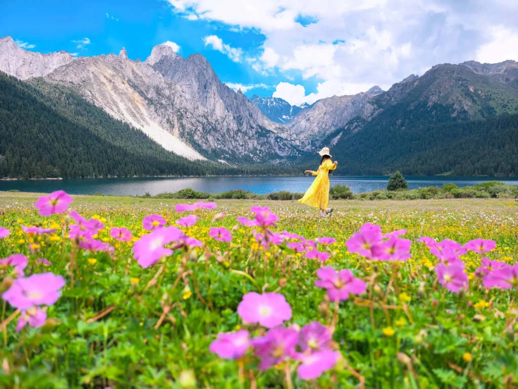 A woman in a bright yellow dress walking through a vibrant field of pink and yellow wildflowers, with a pristine lake and majestic mountains in the background under a blue sky.