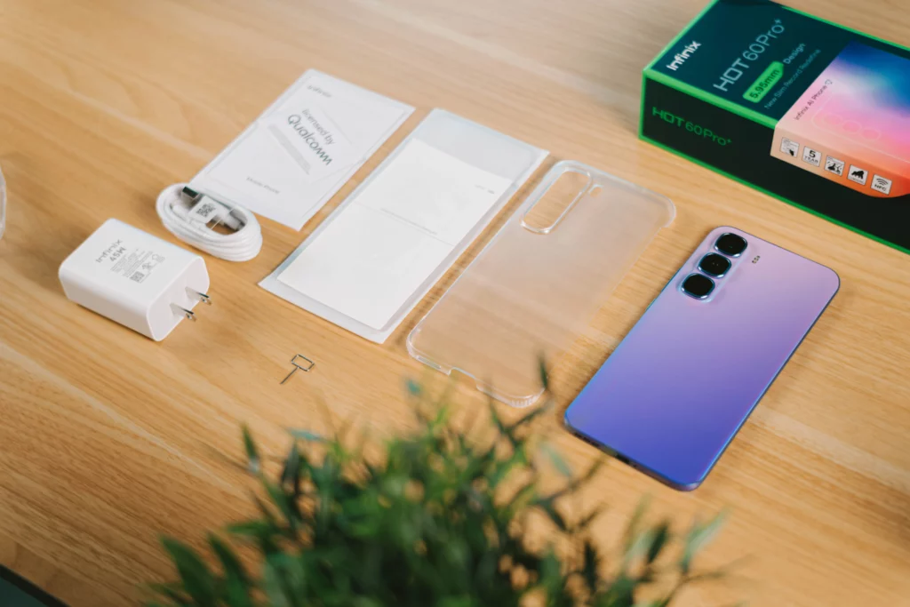 The contents of the Infinix HOT 60 Pro+ box laid out on a wooden table, including the purple smartphone, a clear protective case, a charging brick, a USB cable, and paperwork.