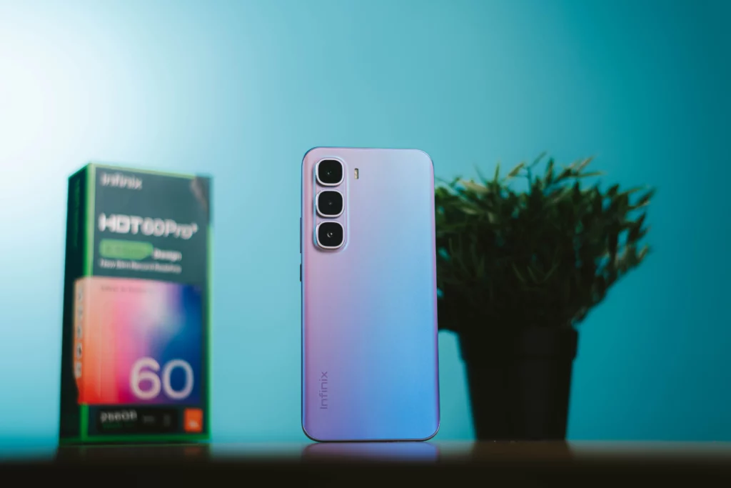 The gradient purple Infinix HOT 60 Pro+ smartphone standing upright next to a potted plant, with its retail box in the background against a blue wall.