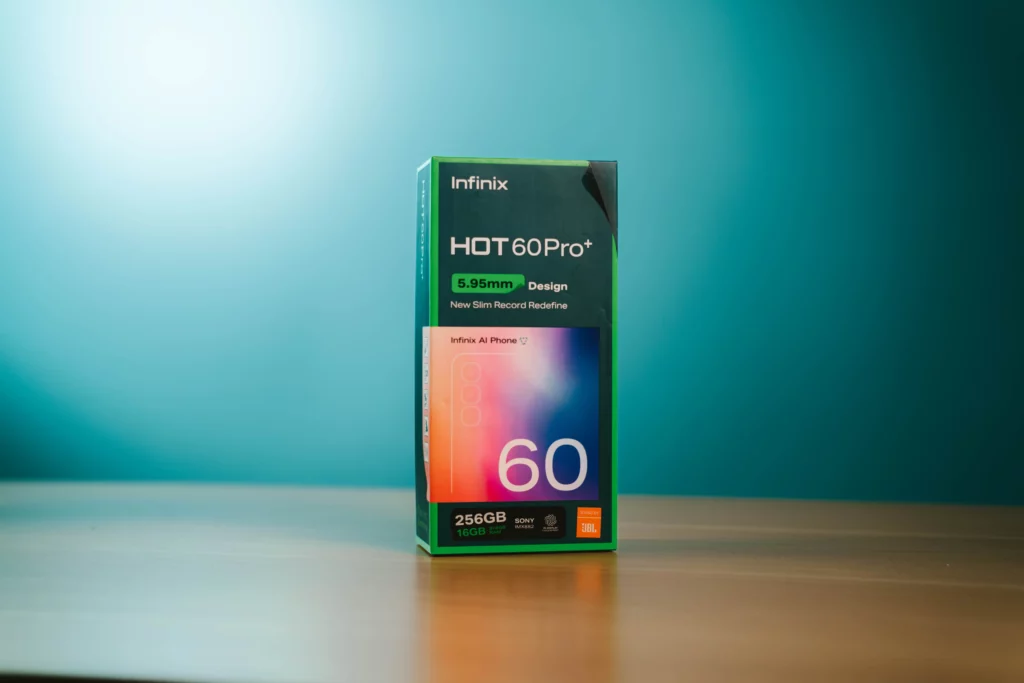 A product shot of the retail box for the Infinix HOT 60 Pro+ smartphone, highlighting key features like "5.95mm Design," "256GB," "16GB RAM," "SONY Camera," and "JBL" on a wooden table.