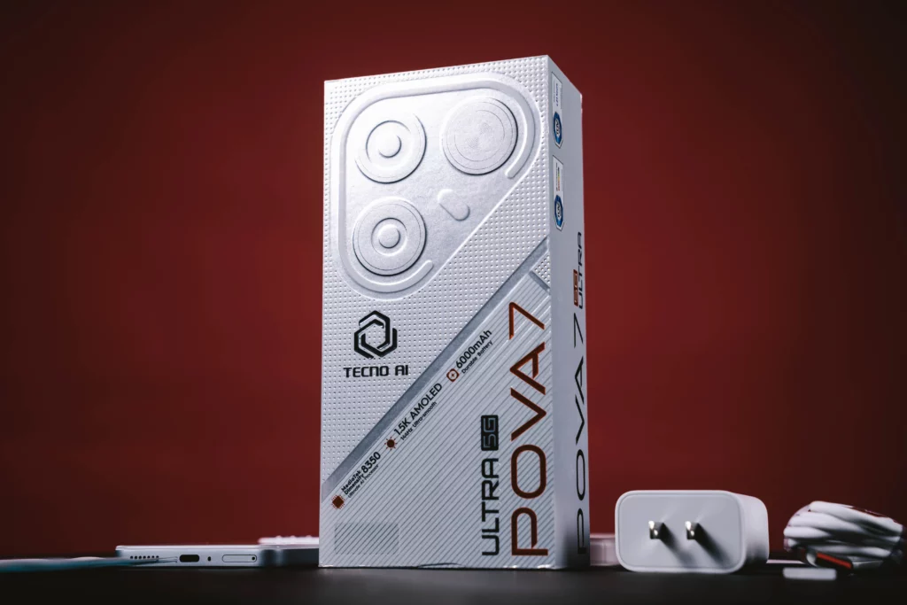 The Tecno POVA 7 Ultra 5G phone box is displayed in an upright position, showing the front design and specifications, with the phone's wall charger and cable visible nearby on a red background.