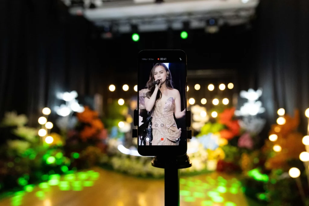 A smartphone, mounted on a stand, is recording a female singer performing on a brightly lit stage. The phone's screen displays a live video feed of the singer, capturing her from a distance. The stage is decorated with numerous lights and floral arrangements.