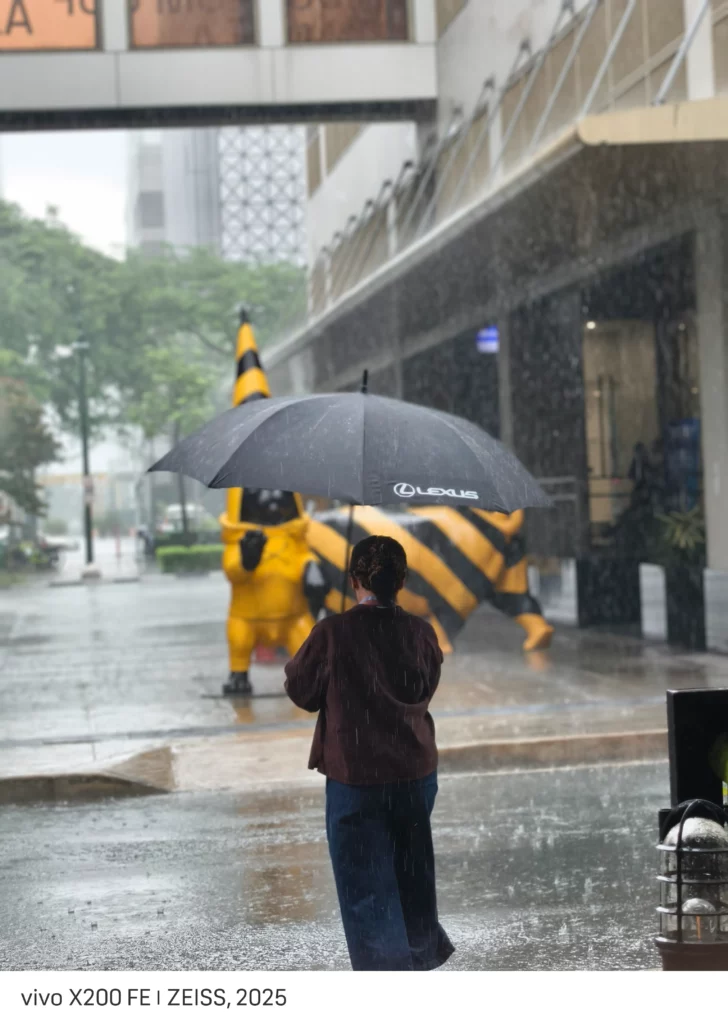 A person holds a black umbrella with a Lexus logo on it while standing in the rain. In the background, two individuals in yellow and black striped costumes are walking. The street is wet from the rain. The text at the bottom reads "vivo X200 FE | ZEISS, 2025".