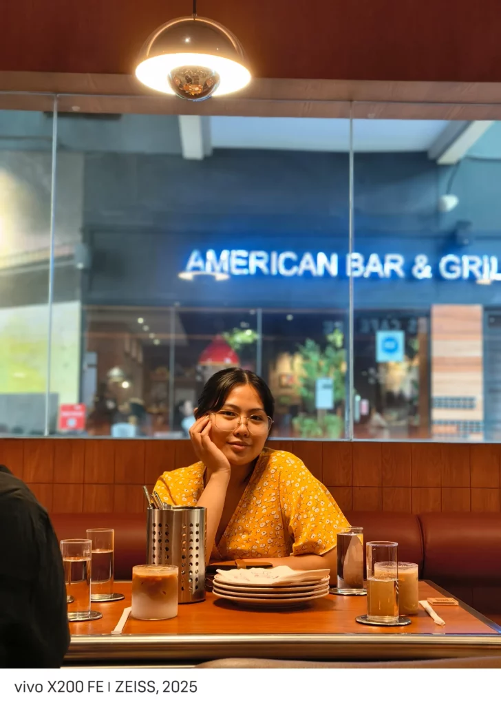 A young woman with glasses, wearing a yellow floral dress, is seated at a restaurant table. She is propping her head up with her hand and smiling slightly. The background shows a sign for "AMERICAN BAR & GRILL" through a large window. The text at the bottom reads "vivo X200 FE | ZEISS, 2025".
