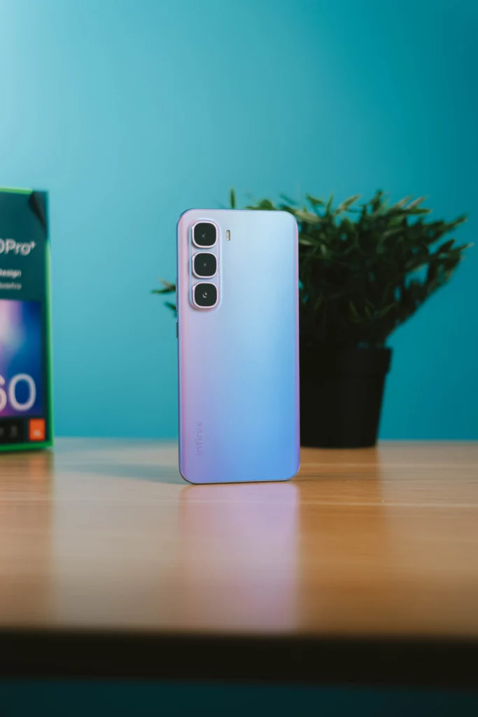 The Infinix HOT 60 Pro+ smartphone, in its unique gradient purple color, standing against a blue background with its retail box and a plant in the background.