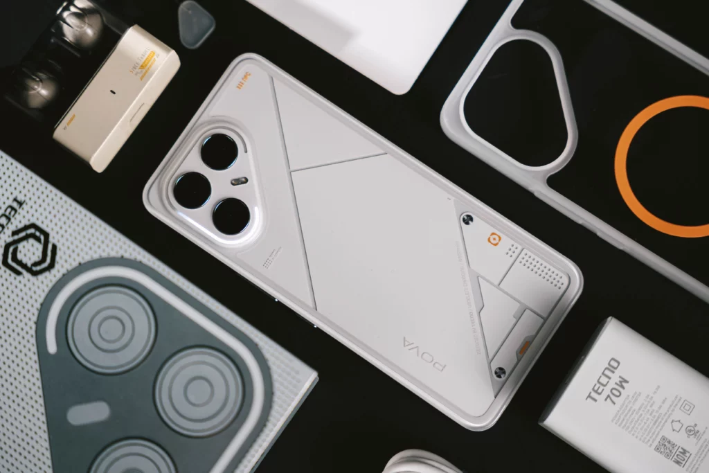 An overhead shot of the Tecno POVA 7 Ultra 5G smartphone, its accessories, and packaging laid out on a dark surface, emphasizing the phone's sleek white design and unique aesthetic.