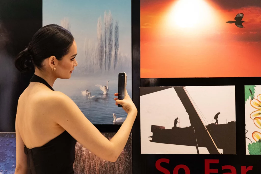 A woman with her hair tied back is holding a golden smartphone up to a large photograph on a wall. She is taking a picture of the photograph, which depicts swans in a body of water. Other photographs, including a silhouette of a bird against a sunset and construction workers, are also visible.