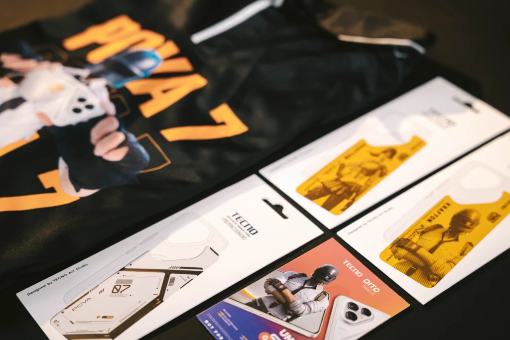 A collection of Tecno and PUBG Mobile promotional items, including a t-shirt and custom phone case packaging featuring character artwork, next to the POVA 7 box.
