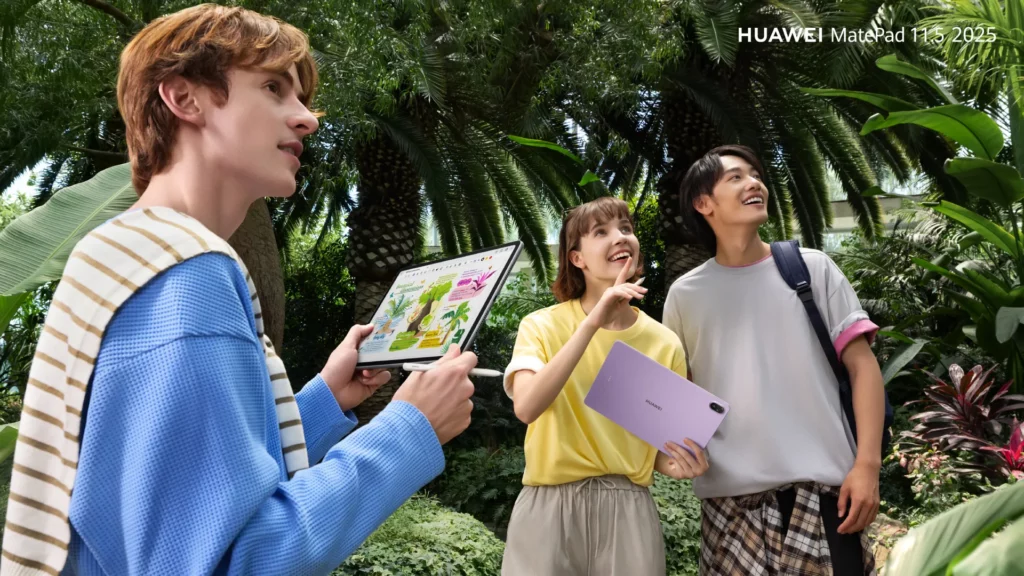 Three friends stand in a lush botanical garden surrounded by tropical plants. One person holds a Huawei MatePad 11.5, showing it to the others as they look up and smile.