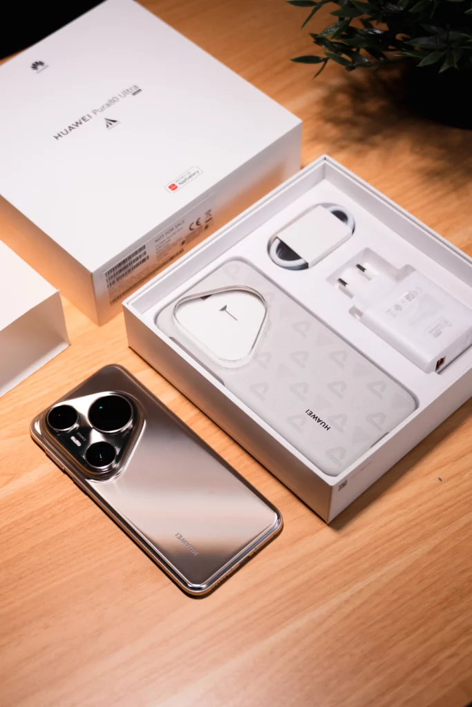 The contents of the HUAWEI Pura80 Ultra box laid out on a wooden table. A silver HUAWEI Pura80 Ultra phone is next to the open box, which contains a clear phone case, a charging brick, and a USB-C cable.