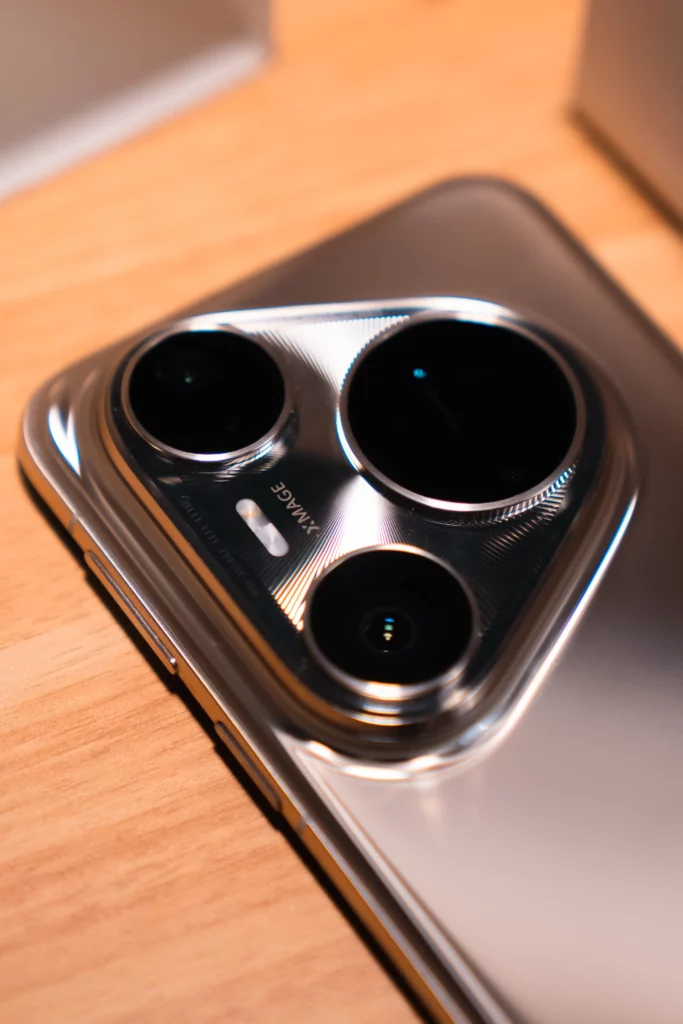 A detailed, angled close-up of the rear camera module of a silver HUAWEI Pura80 Ultra smartphone, highlighting the three large lenses and the "XMAGE" branding. The phone lies on a wooden surface.