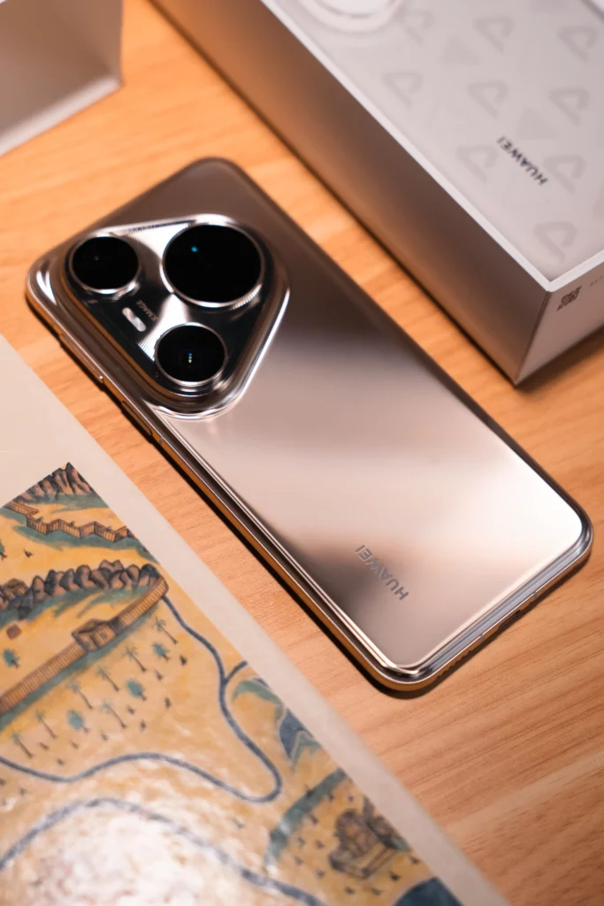 A silver HUAWEI Pura80 Ultra smartphone rests face down on a wooden table, with its camera module facing upwards, next to an open phone box and a book with illustrations.