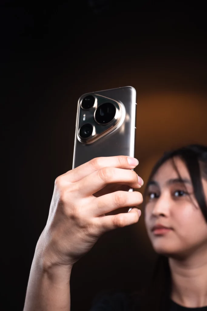 A hand holds a silver HUAWEI Pura80 Ultra smartphone, showing its prominent camera module, with a person's blurred face in the background looking away from the camera.