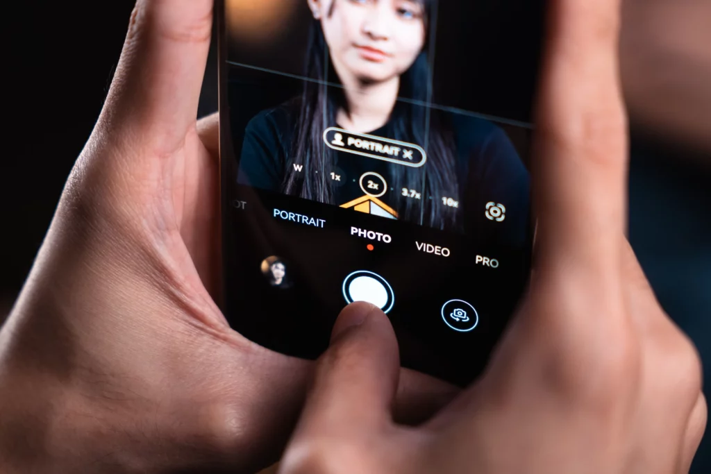 A close-up of hands holding a smartphone, with a finger about to tap the shutter button in the camera app interface. The screen shows a person's face in "Portrait X" mode, with zoom options (1x, 2x, 3.7x, 10x) and modes like "PORTRAIT," "PHOTO," "VIDEO," and "PRO."