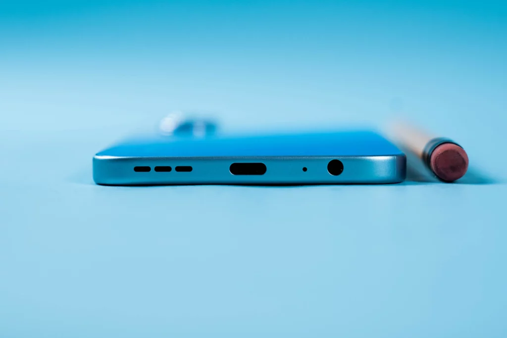 A macro shot of the bottom edge of a light blue Infinix smartphone on a matching light blue surface. The ports and speaker grills are clearly visible, along with a pencil with an eraser resting nearby.