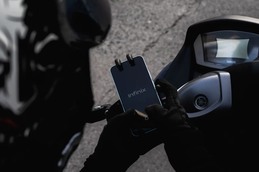 A person wearing black gloves holds a black smartphone with the Infinix logo visible on the screen. The smartphone is attached to a mount, and in the blurred background, parts of a motorcycle's dashboard and handlebars are visible.
