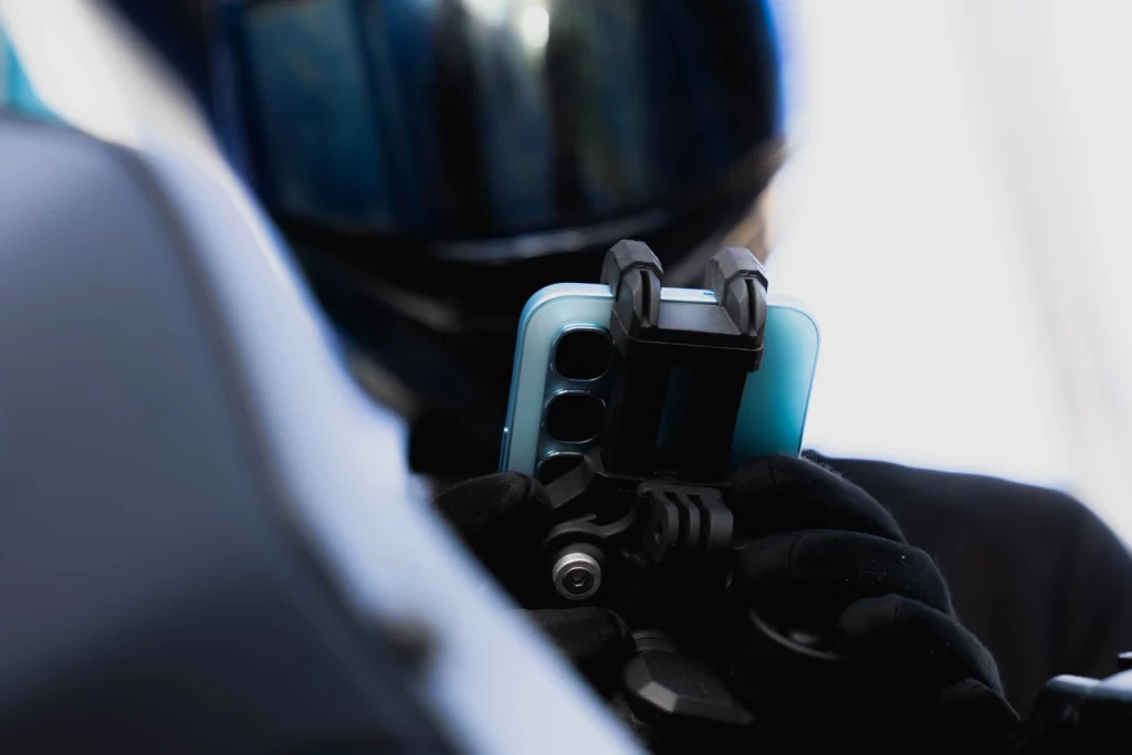 A close-up shot of a light blue smartphone being attached to a black phone mount. A person wearing a helmet and black gloves is holding the device. The phone's triple-camera system is visible on the back.