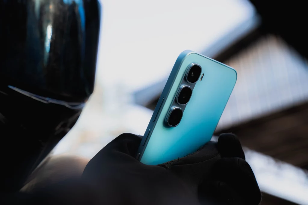A close-up, low-angle shot of a person in black gloves holding a light blue smartphone. The phone's three-camera system is prominent on the back, and the person's helmet is partially visible in the left foreground. The background is a bright, blurry outdoor setting.