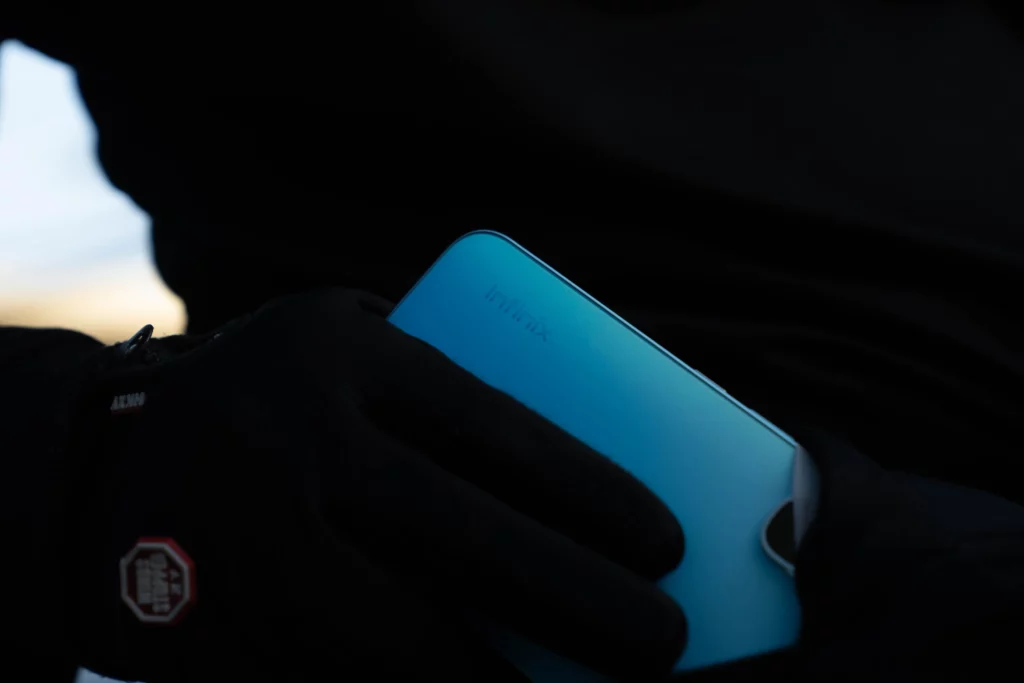 A low-angle, close-up shot of the back of a light blue smartphone with the "Infinix" logo visible. The phone is held by two gloved hands, and the lighting is dark, with the background and the person's body in silhouette.