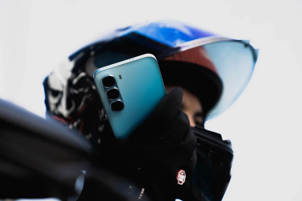 A medium shot of a person in a helmet with a tinted visor holding a light blue smartphone. The phone's triple-camera setup is clearly visible. The person is wearing black gloves, and the background is a bright, out-of-focus sky.