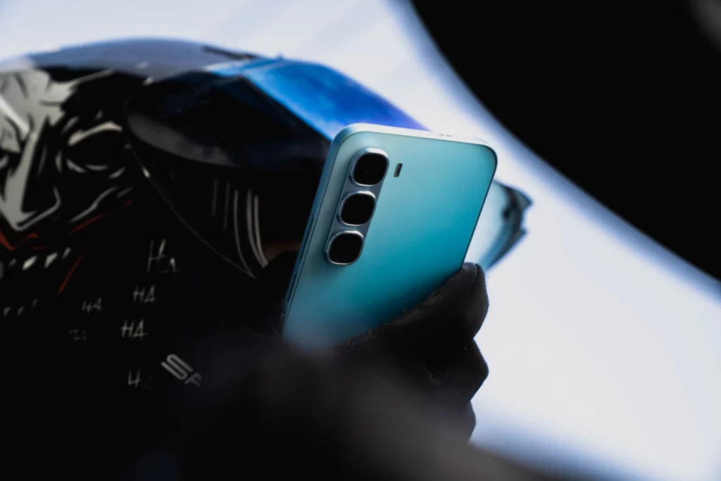 A close-up shot of a person wearing a helmet and black gloves, holding a light blue smartphone. The phone's camera module with three lenses is visible on the back. The background is bright and out of focus.