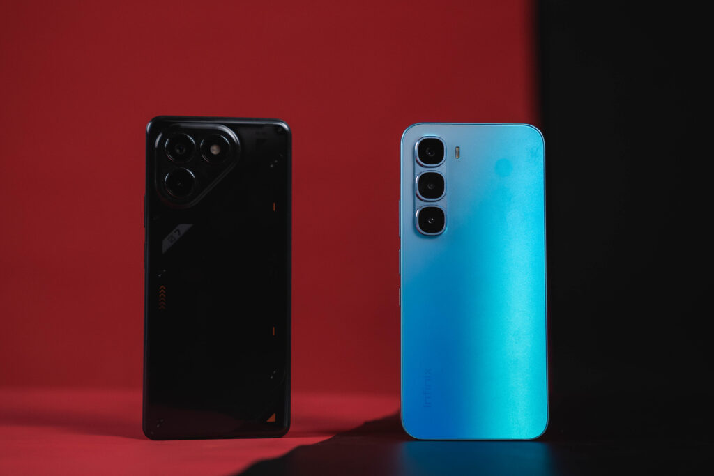 A product shot of two smartphones placed side-by-side against a red and black background. The phone on the left is black with a stylized, mechanical back panel and a large, circular camera module. The phone on the right is a vibrant blue with a smooth back and a vertical triple-camera setup.