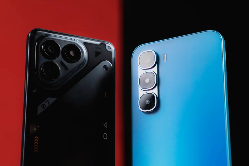 A close-up shot focusing on the rear camera modules of two smartphones. The left phone, in black, features a large, distinct camera island with three lenses and visible screw details, giving it a mechanical look. The right phone, in blue, has a simpler, vertical stack of three cameras. The background is split between black and red.