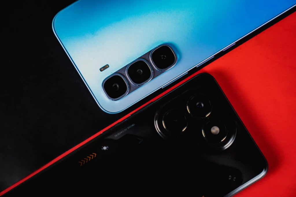A close-up, diagonal shot of the top backs of two smartphones against a red and black background. The blue phone is in the foreground, showing its vertical triple-camera setup. The black phone is in the background, showing its more elaborate, mechanical-style circular camera island with three lenses.